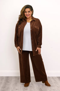 Front view of a plus sized female model wearing a brown button-up corduroy jacket with flap pockets and matching loose-fit pants, against white wall and gray floor