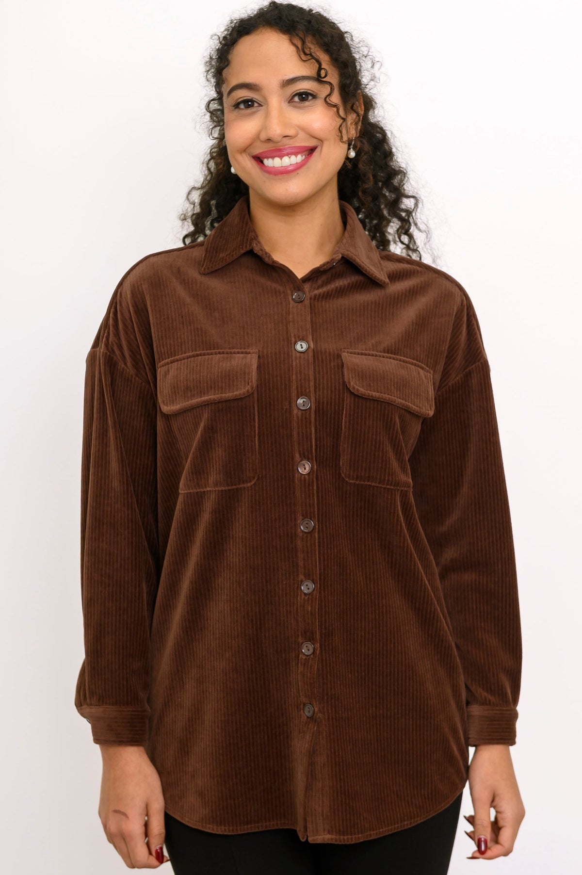 Front view of a female model wearing a dark brown button-up corduroy shirt jacket with double flap pockets, with black pants, against a white wall and gray floor
