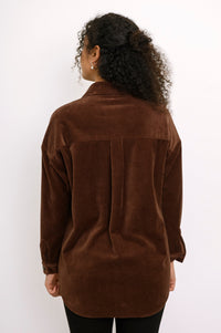 Back view of a female model wearing a dark brown corduroy jacket featuring a pleated back yoke and curved hem, against white wall and gray floor