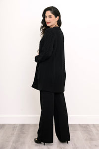 Side and rear view of a woman wearing a hip length black cable knit cardigan showing clean shoulder tailoring.