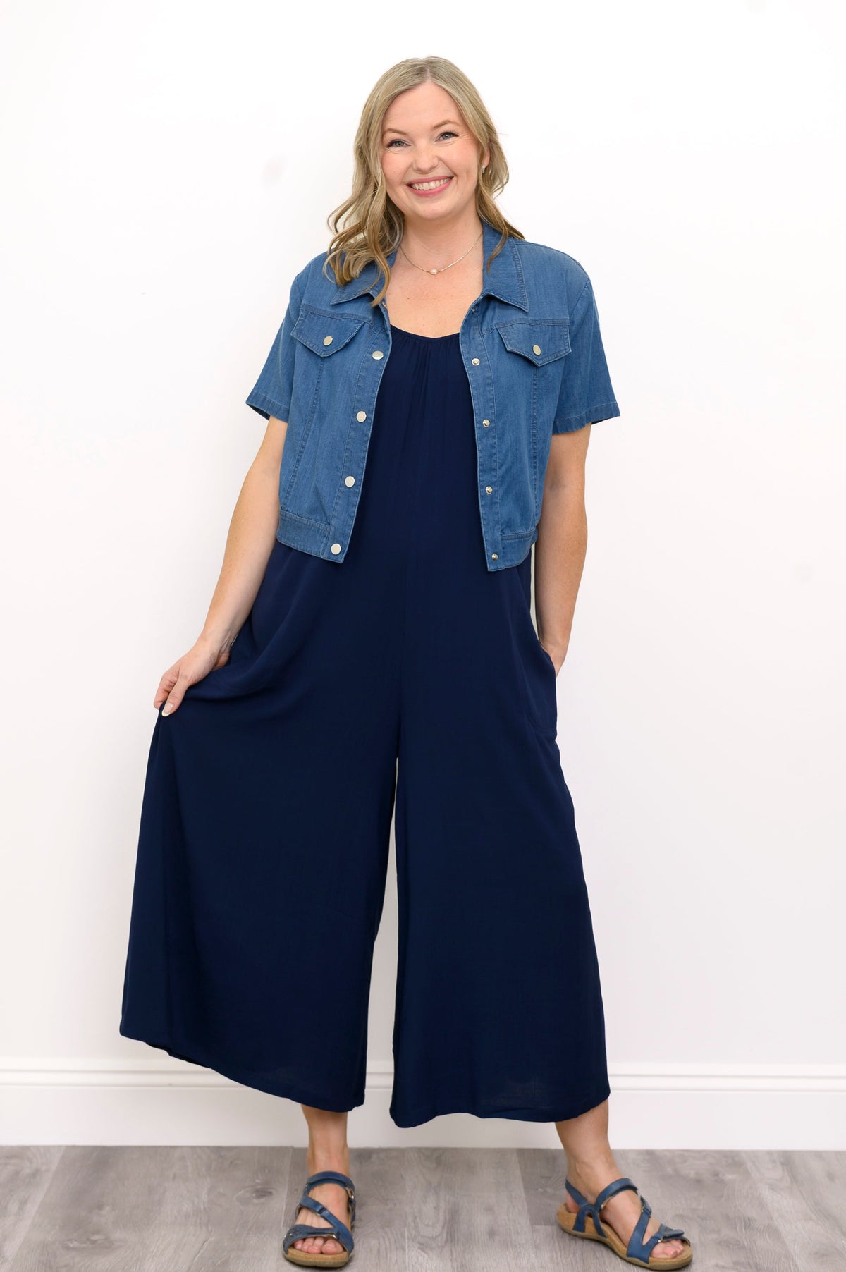 Front view, woman wearing blue jacket, navy blue jumpsuit, and sandals against a white background