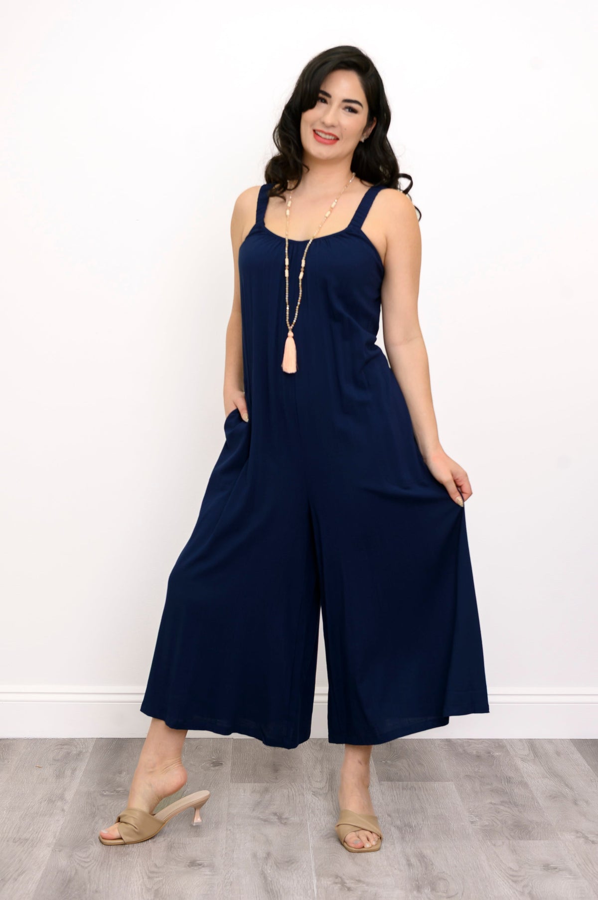 Woman wearing a navy blue jumpsuit, tassel necklace, and beige sandals against a white background, front view