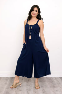 Woman wearing a navy blue jumpsuit, tassel necklace, and beige sandals against a white background, front view