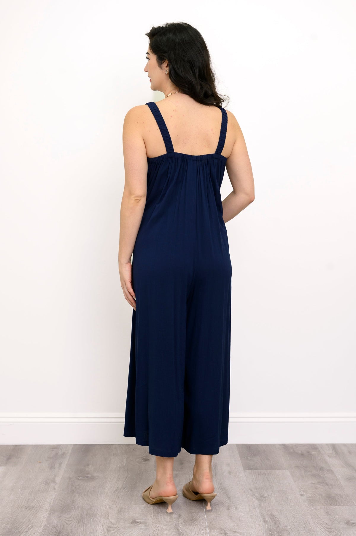 Woman wearing a navy blue jumpsuit and beige sandals against a white background, back view