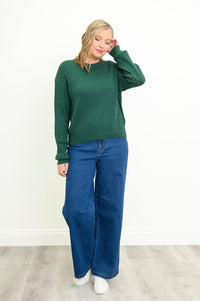 Front view of a female model wearing a green sweater with denim pants and white shoes, tucking hair behind ear, against a white wall and gray floor
