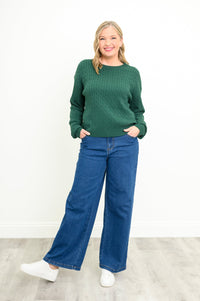 Front view of a female model wearing a green sweater with denim pants and white shoes, against a white wall and gray floor