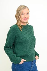 Side-facing view of a female model wearing a green sweater with denim pants, hands in pockets, against a white backdrop