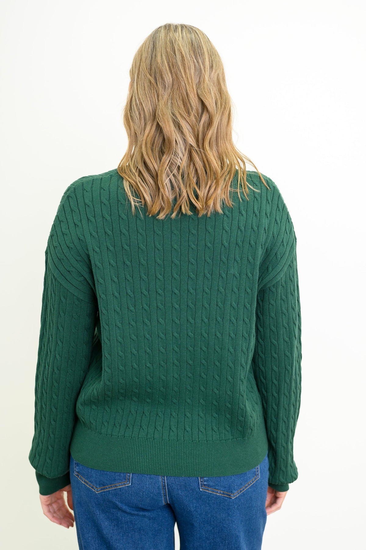 Back view of a a female model wearing a green sweater with denim pants, against a white backdrop