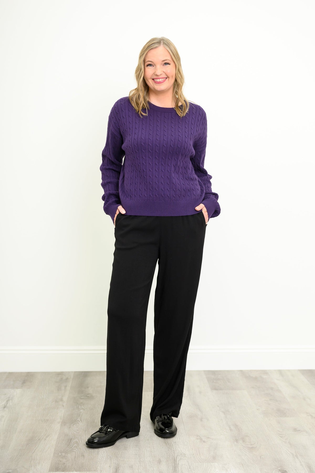 Front view of a female model wearing a purple sweater with black pants and shoes, against a white wall and gray floor