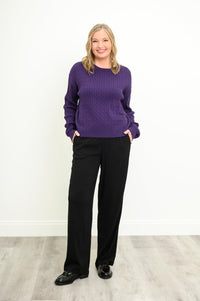 Front view of a female model wearing a purple sweater with black pants and shoes, against a white wall and gray floor