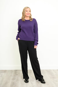 Front view of a female model wearing a purple sweater with black pants and shoes, with hand in pocket, against a white wall and gray floor