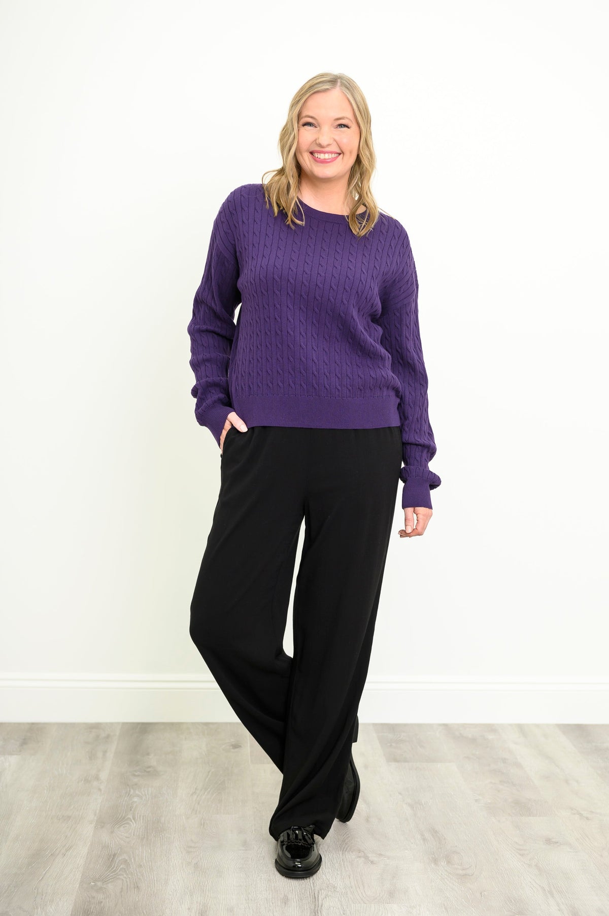 Front view of a female model wearing a purple sweater with black pants and shoes, with one propped leg, against a white wall and gray floor