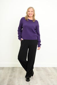 Front view of a female model wearing a purple sweater with black pants and shoes, with one propped leg, against a white wall and gray floor