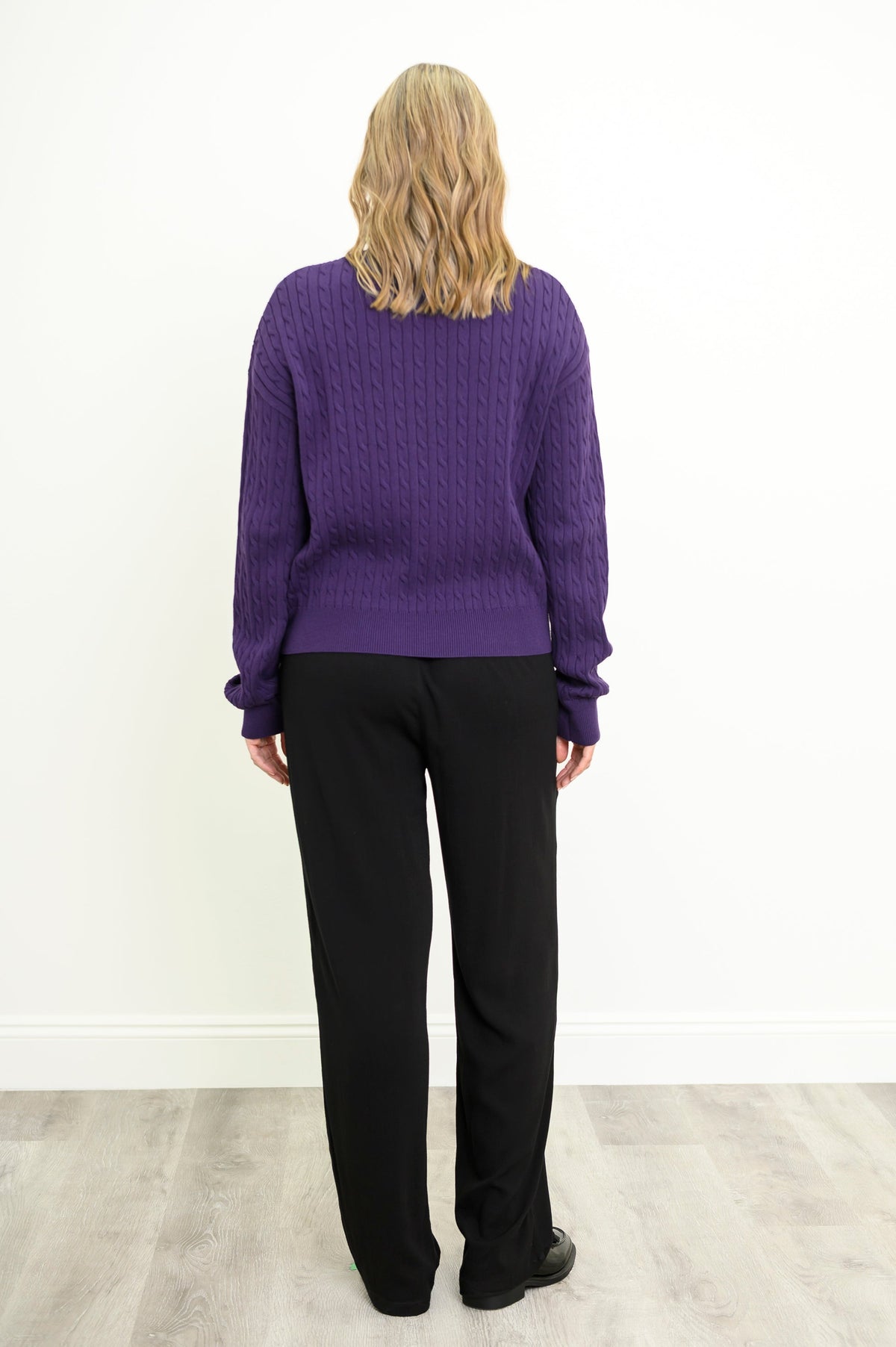 Rear view of a female model wearing a purple sweater with black pants and shoes, against a white wall and gray floor