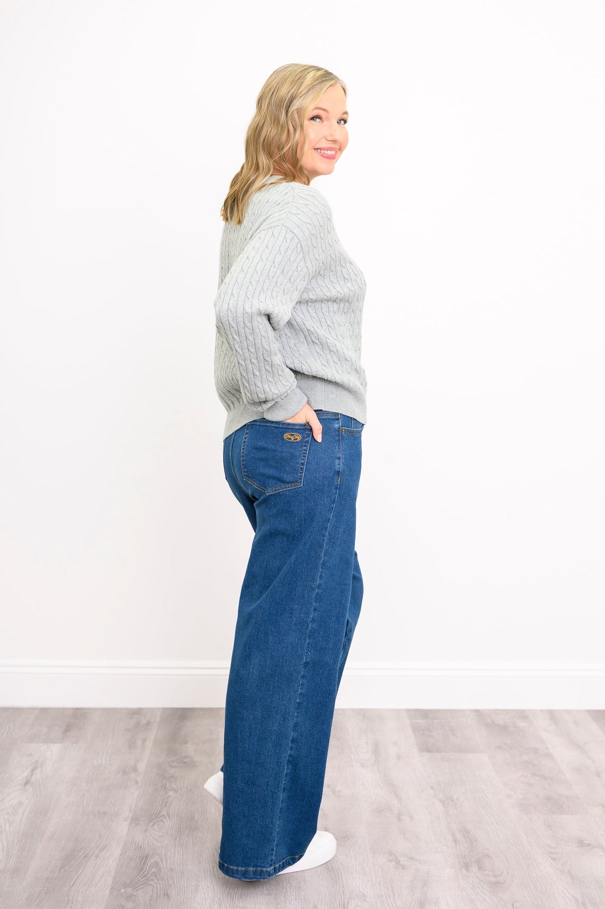 Side view of a female model wearing a light grey sweater with denim pants and white shoes, against a white wall and gray floor