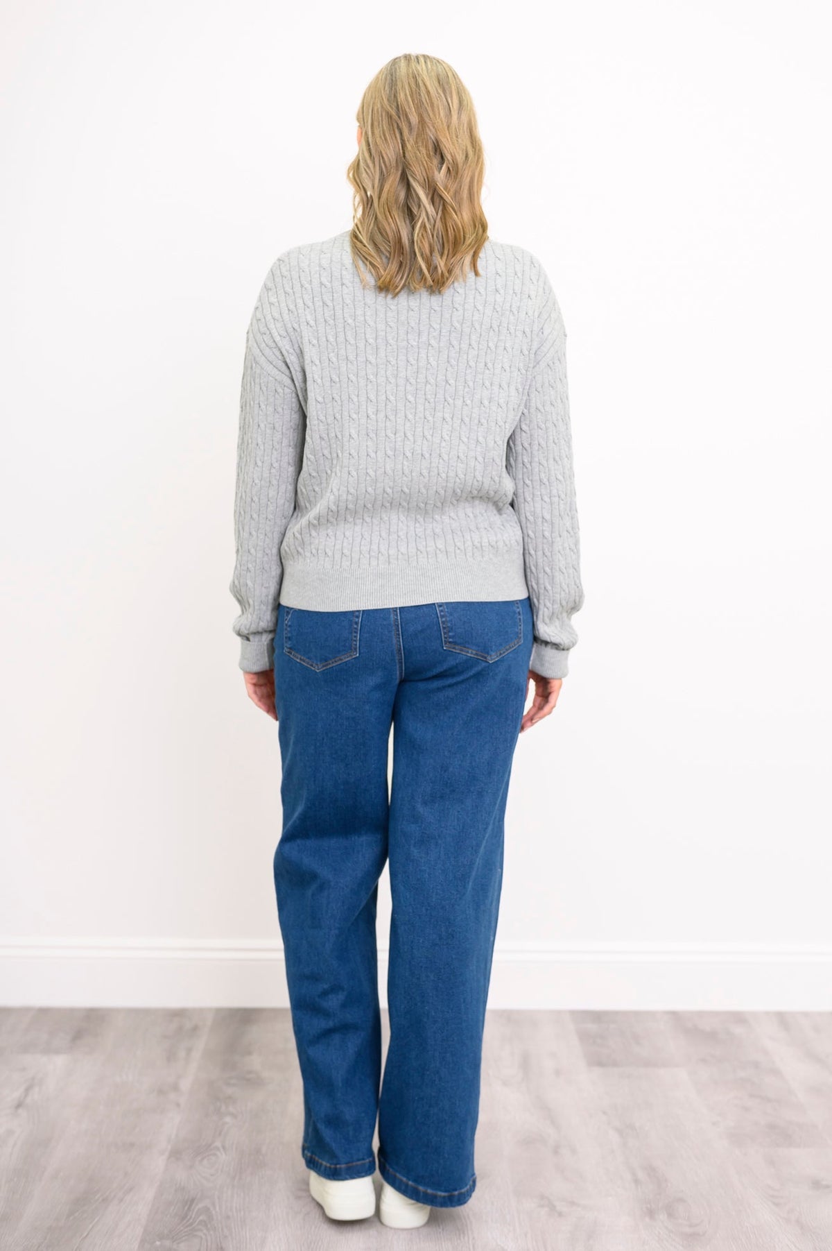 Back view of a female model wearing a light grey sweater with denim pants and white shoes, against a white wall and gray floor