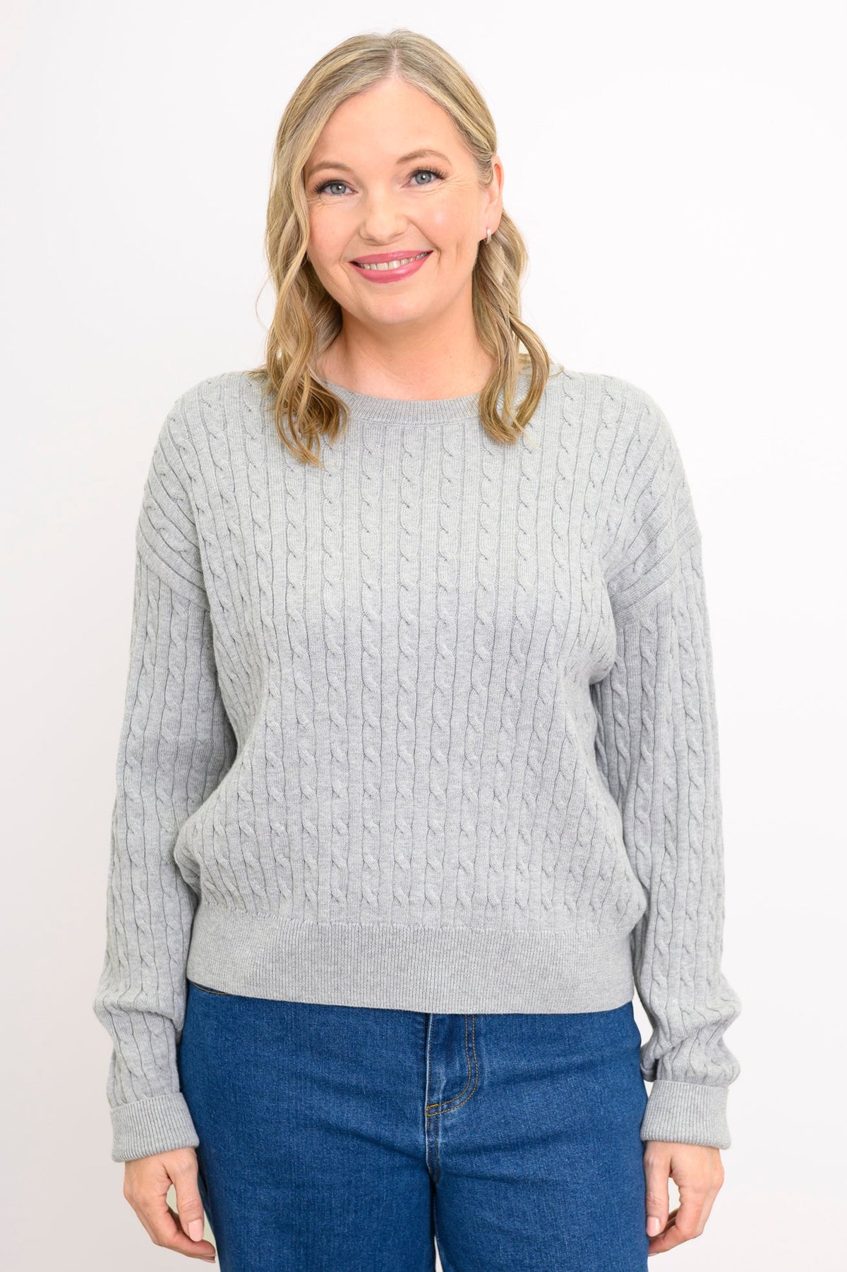 Front view of a female model wearing a light grey sweater with a round neck, styled with denim pants against white backdrop