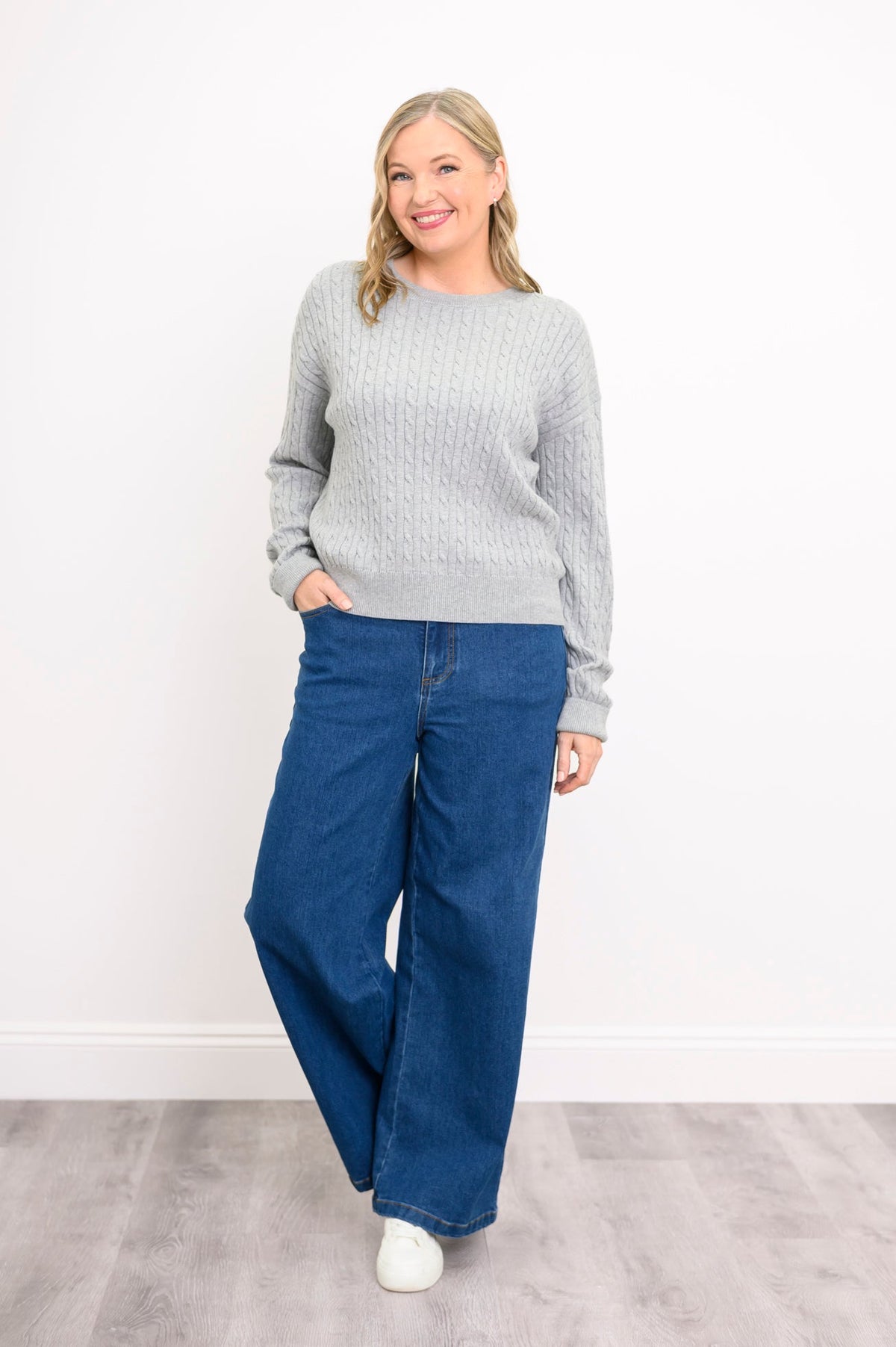 Front view of a female model wearing a light grey sweater with denim pants and white shoes in relaxed pose, against white wall and gray floor