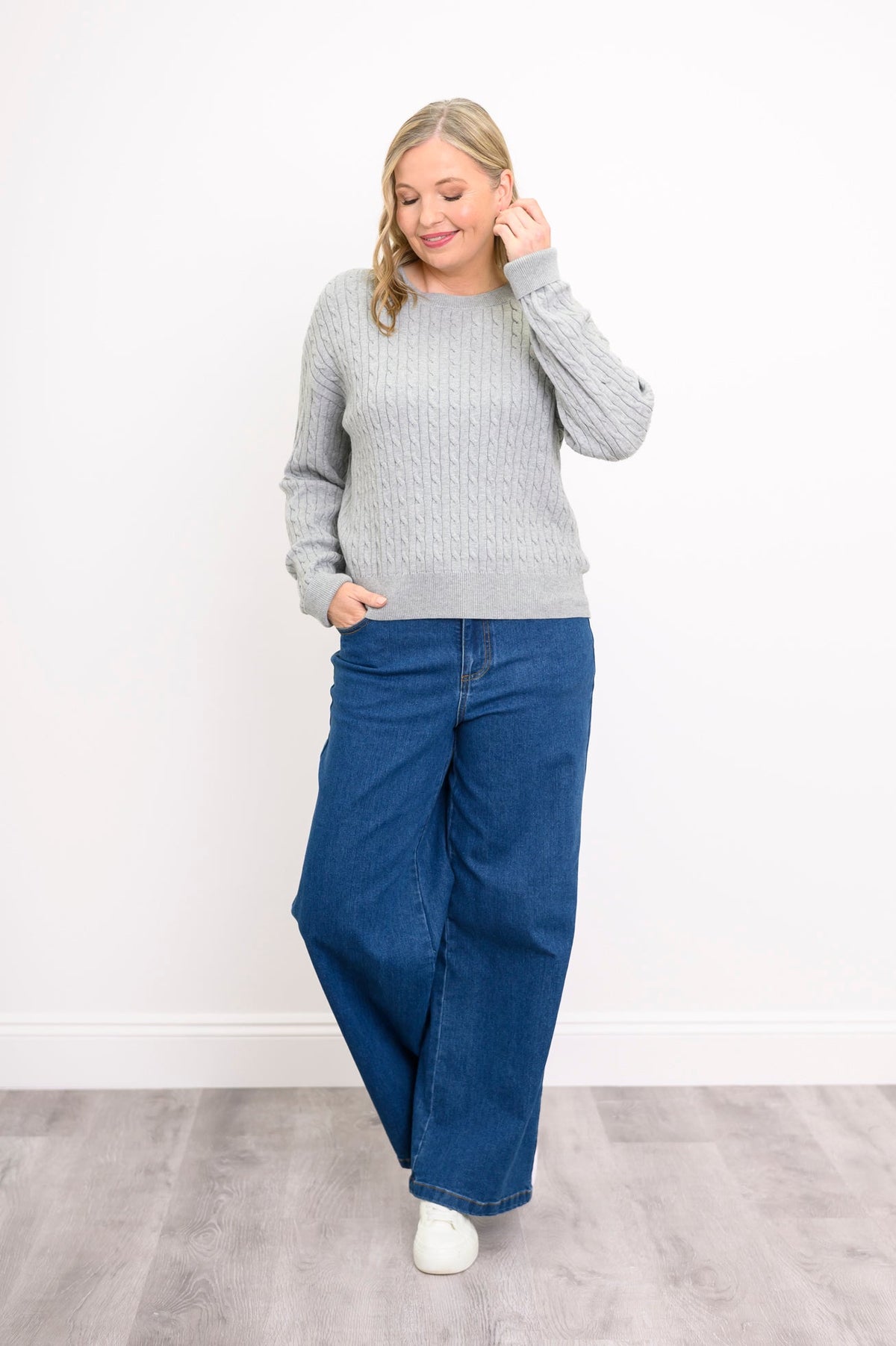 Front view of a female model wearing a light grey sweater with denim pants and white shoes, tucking hair behind ear, against white wall and gray floor