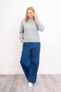 Front view of a female model wearing a light grey sweater with denim pants and white shoes, tucking hair behind ear, against white wall and gray floor
