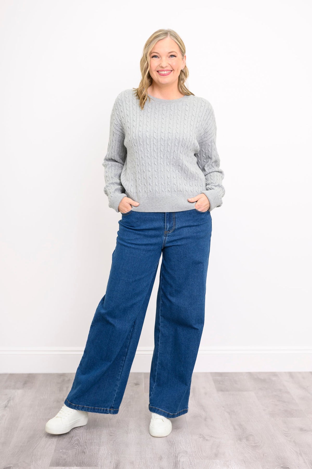 Front view of female model wearing a light grey sweater with denim pants and white shoes, against white wall and gray floor