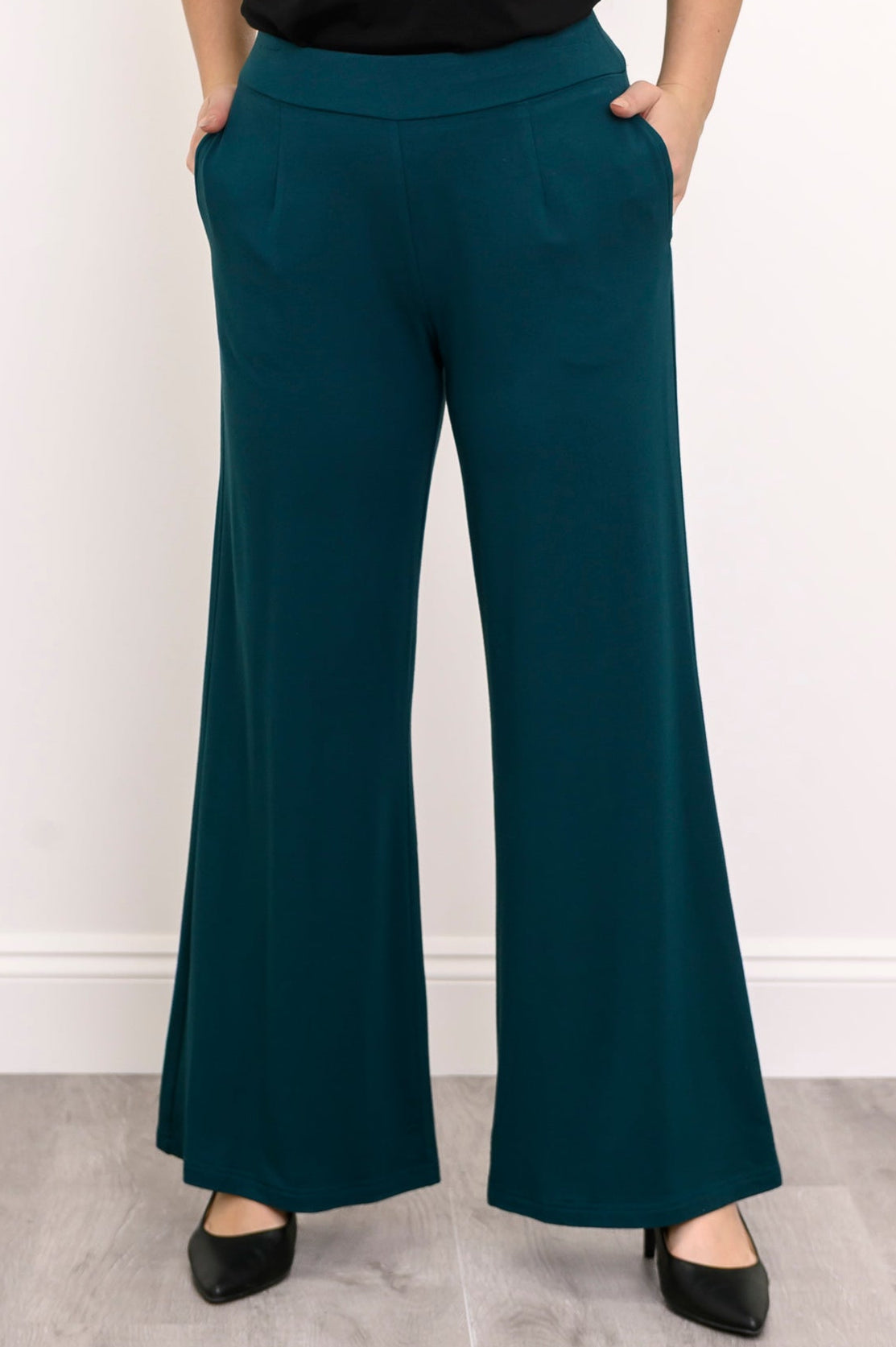 Close-up front view of a female model wearing teal wide-leg French Terry pants with hands in side pockets with black heels, against a white background and grey floor
