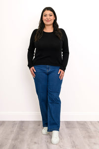 Front view of a plus size female model wearing a black sweater with a flat finished hemline, with denim pants, against a white wall and gray floor
