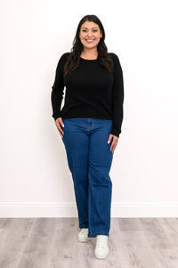 Front view of plus size female model in a black sweater featuring extended coverage sleeves, styled with casual blue denim pants, against a white wall and gray floor