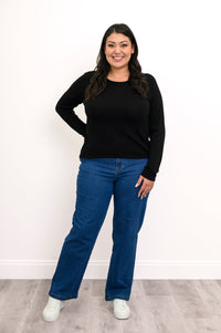 Front view of plus size female model wearing a black long-sleeve sweater featuring clean flat-finished wrist cuffs, hand on hip, with denim pants and white shoes. Against a white wall and gray floor
