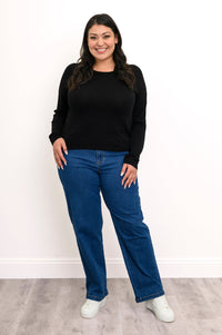 Front view of a plus size female model wearing a black sweater, showcasing a straight-cut hemline, with denim pants and white shoes, against a white wall and gray floor
