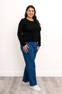 Side-facing view of a plus size female model wearing a black sweater with denim pants and white shoes, against a white wall and gray floor