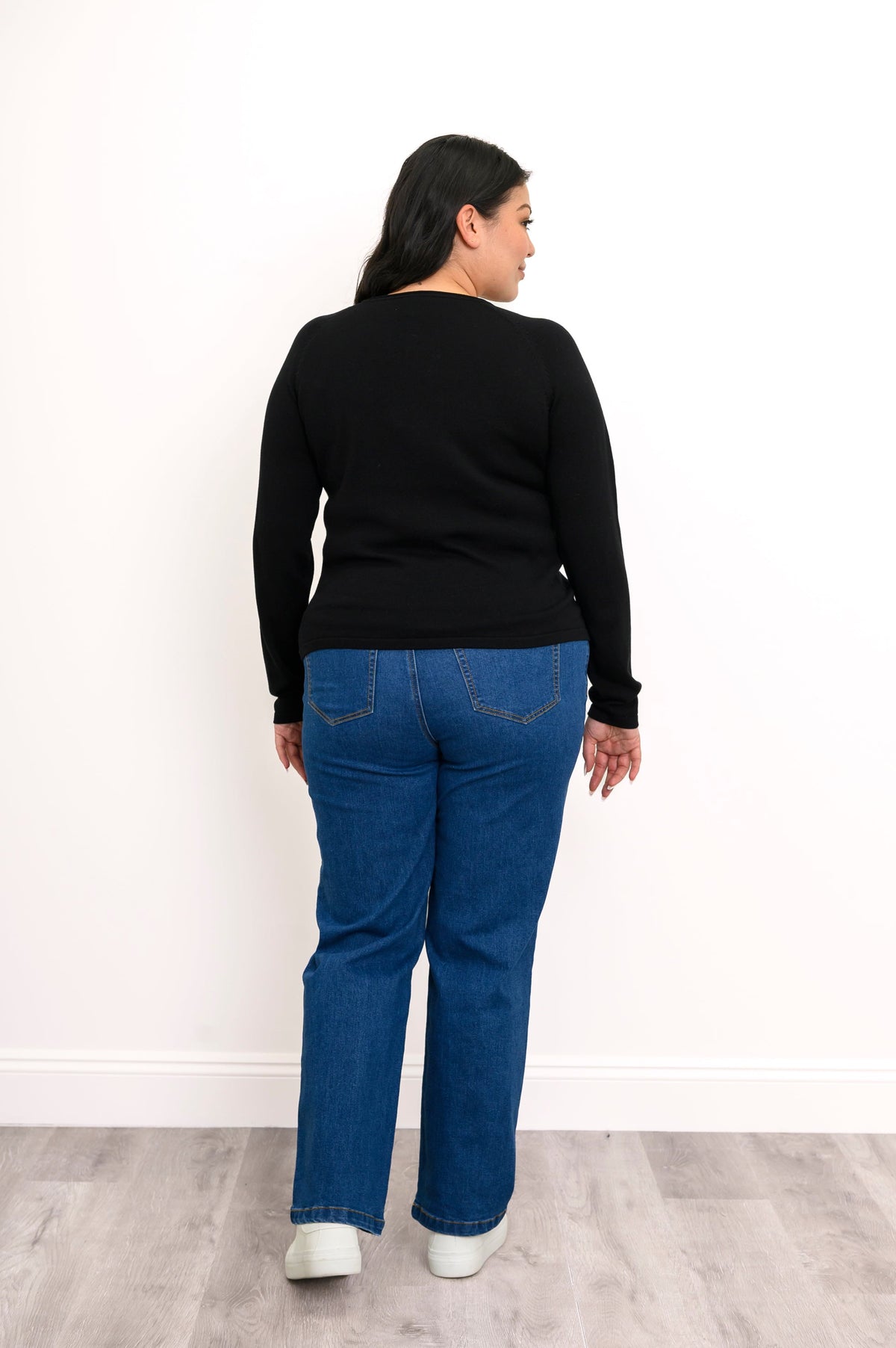 Back view of a plus size female model wearing a black sweater showcasing a seamless rear panel, long sleeves, with classic blue denim jeans and white shoes. Against a white wall and gray floor
