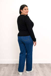 Back view of a plus size female model wearing a black sweater, with denim pants and white shoes, on a white wall and gray floor
