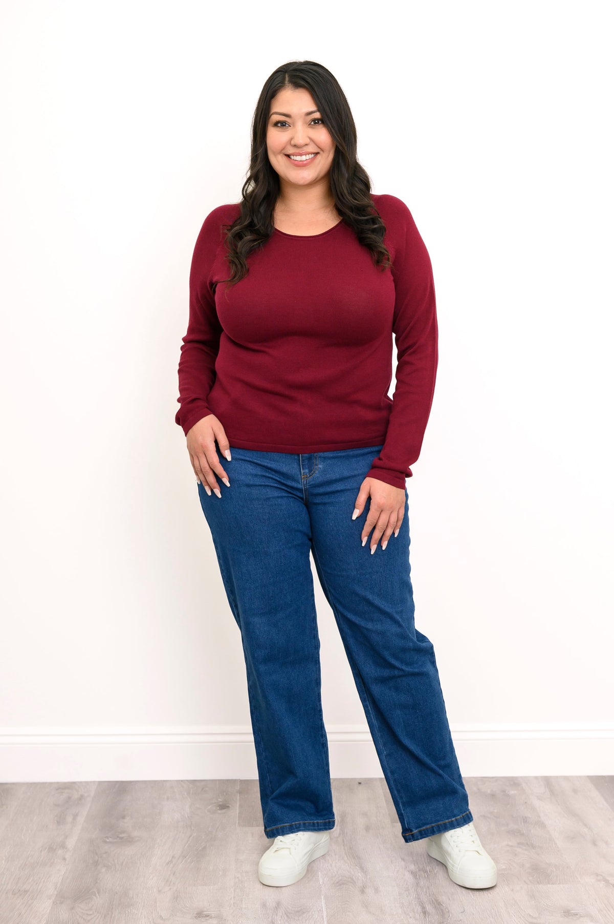 Front view of plus sized female model wearing a wine red sweater featuring a flat finished hemline, styled simply with denim pants and white shoes. Against white wall and gray floor