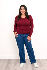 Front view of plus sized female model wearing a wine red sweater featuring a flat finished hemline, styled simply with denim pants and white shoes. Against white wall and gray floor