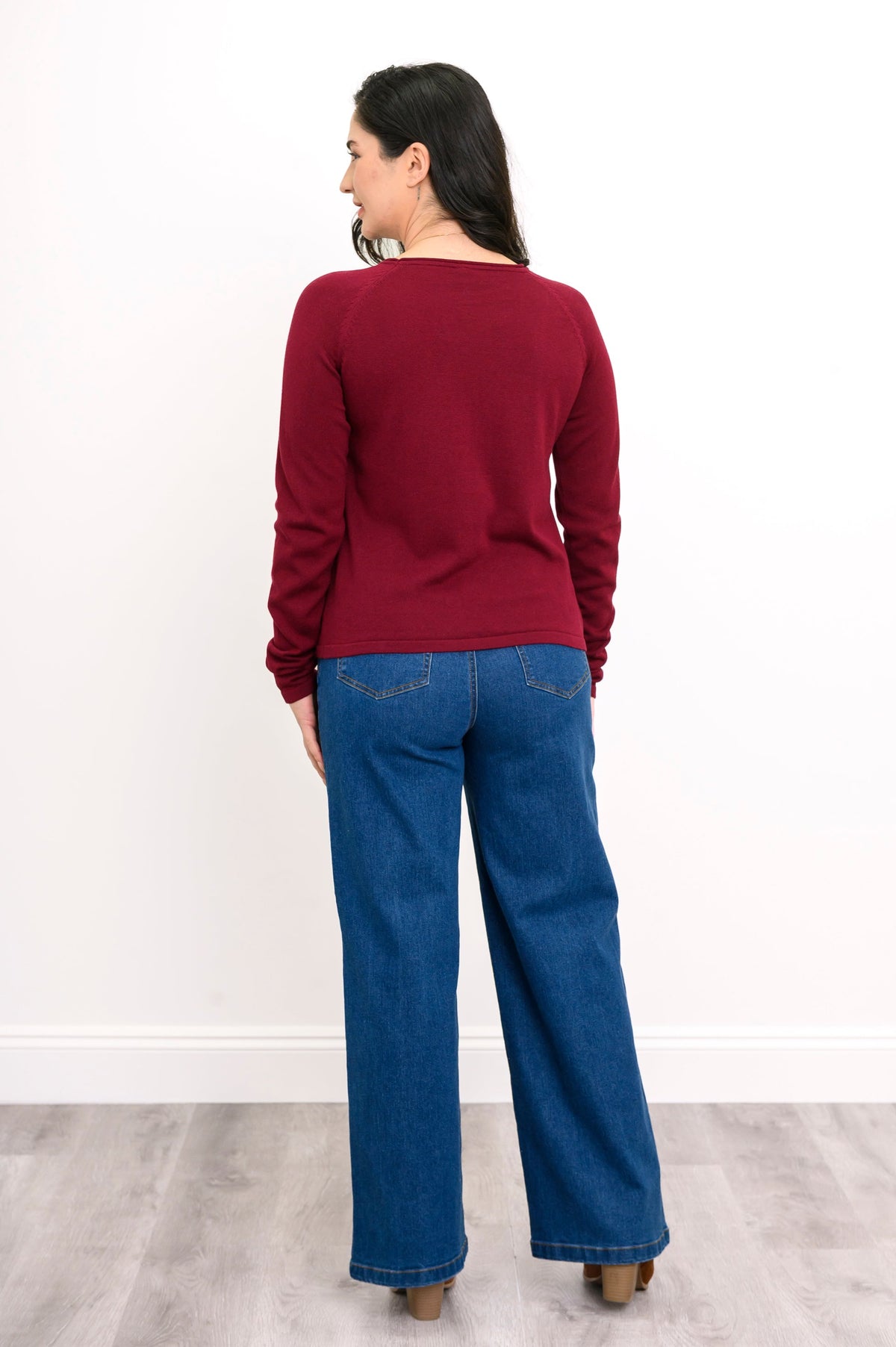 Back view of a female model wearing a long-sleeve wine red sweater, styled with blue jeans and brown shoes against a white wall and gray floor