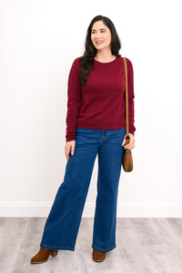 Front view of a female model wearing a wine red sweater and denim jeans with a over-shoulder handbag and brown shoes. Against a white wall and gray floor