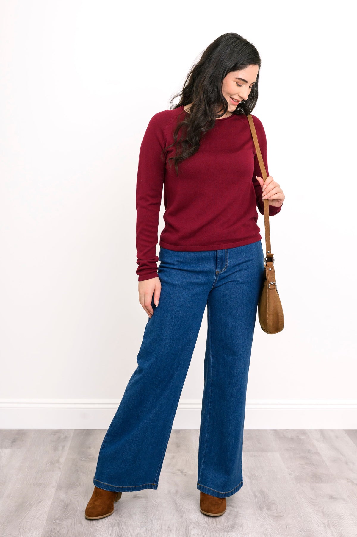 Front view of female model wearing a wine-red sweater layer and blue denim pants with handbag and brown shoes. Against white wall and gray floor