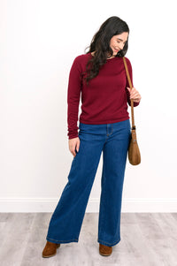 Front view of female model wearing a wine-red sweater layer and blue denim pants with handbag and brown shoes. Against white wall and gray floor