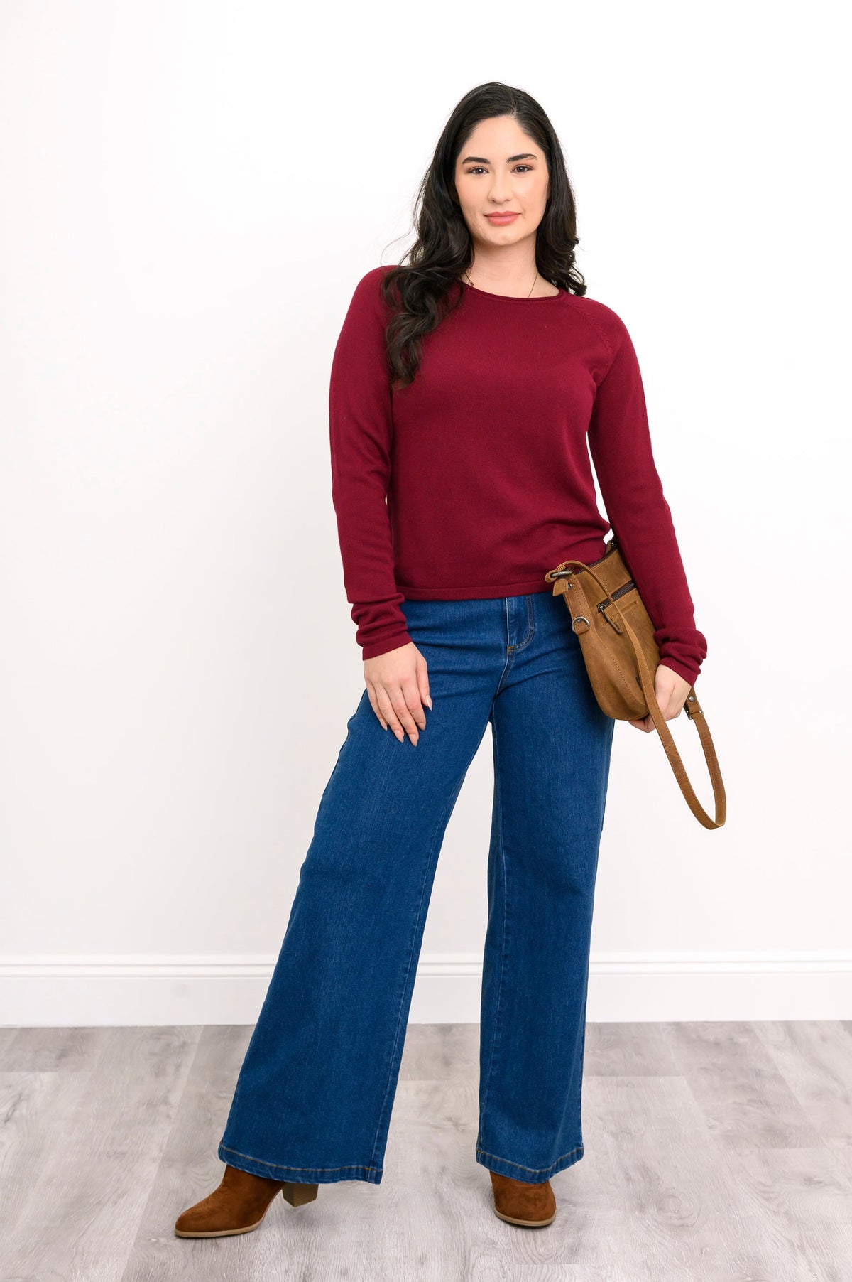 Front view of female model wearing wine red sweater with long raglan sleeves and a rounded hem, with denim pants and a handbag, against a white wall.
