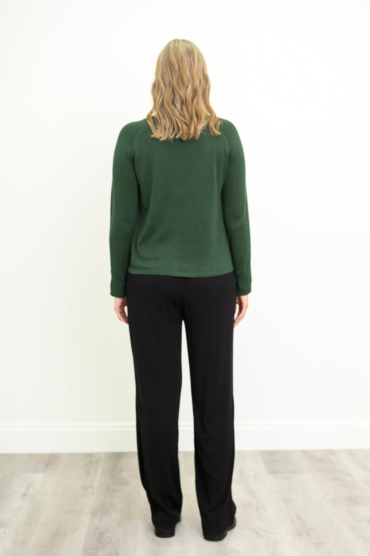 Back view of a female model wearing a green sweater, styled with black pants and shoes, against a white wall and gray floor