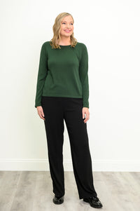 Front view of a female model wearing a green sweater and slim black pants, with black shoes, against a white wall and gray floor