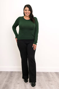 Front view of a plus size female model wearing a green sweater with black pants and shoes, against white wall and gray floor