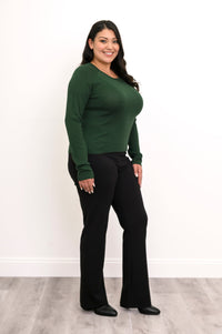 Side-facing view of a plus size female model wearing a green sweater with black pants and shoes, against a white wall and gray floor