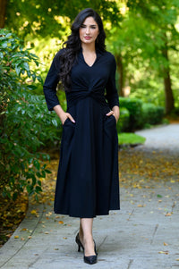 front view of female model wearing a black midi wrap dress with three-quarter sleeves with hands in side pockets in a green park