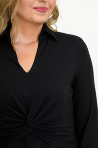 Close up view of female model wearing a black midi dress with three-quarter sleeves, a V-neckline, and a twisted waist detail against a white wall