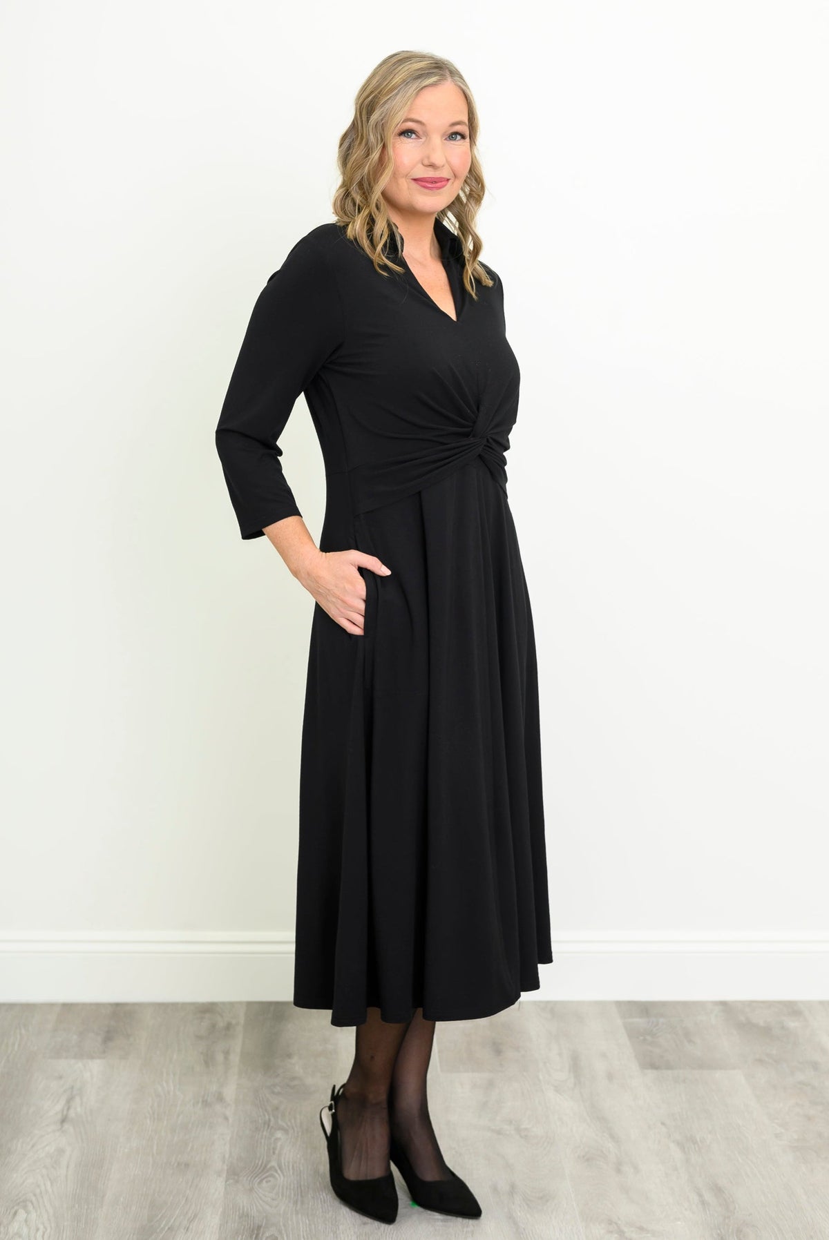 front view of female model wearing a black midi dress with three-quarter sleeves, a V-neckline, and a twisted waist detail, standing against a white wall and light gray wood floor.