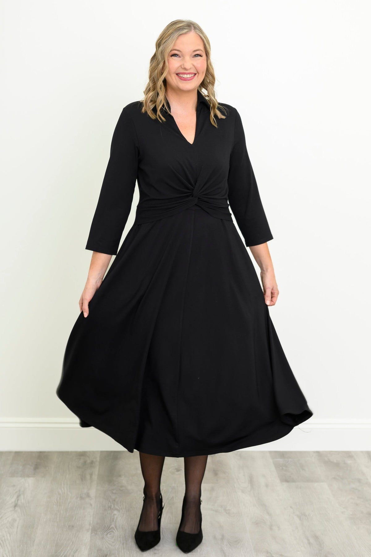 front view of female model wearing a black midi dress with three-quarter sleeves, a V-neckline, and a twisted waist detail, holding out the dress against a white wall and light gray wood floor.