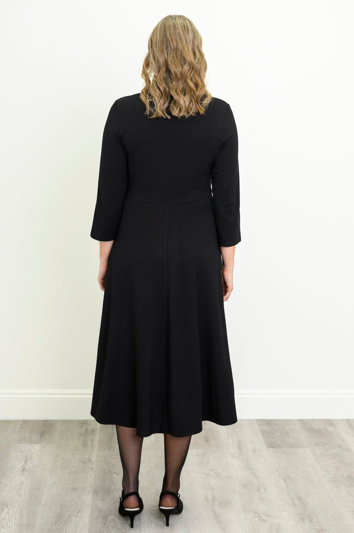 back view of female model wearing a black midi dress with three-quarter sleeves, a V-neckline, and a twisted waist detail, standing against a white wall and light gray wood floor.
