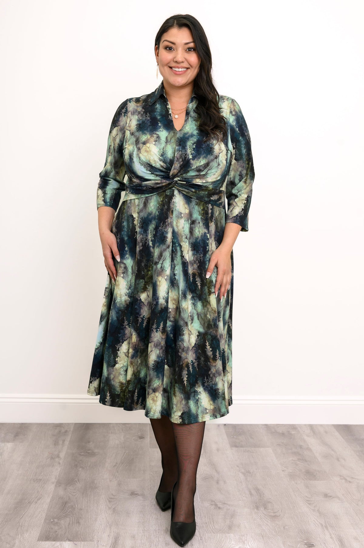 Front view of plus sized female model wearing a flowy bamboo A-line dress featuring a shirt collar, V-neck, and defined high waist twist, against a white wall and gray floor